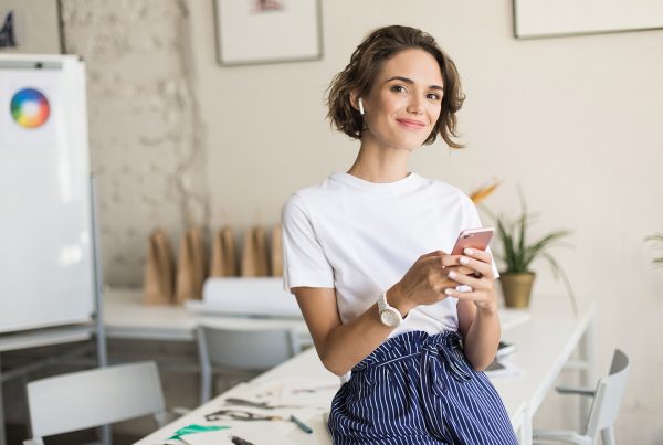 Jóven mujer escribiendo un post como comunity manager desde un teléfono móvil sentada sobre la mesa de su oficina
